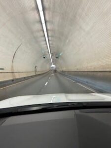 View of the Mobile Bay Tunnel through the windshield of a vehicle. The tunnel is covered in white tiles and has a row of lights running down the center of the highest point on the ceiling. You can see daylight at the far end of the tunnel.