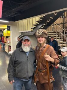 Two individuals posing for a photo, one wearing a gray fleece jacket and the other in a historical costume with a fur hat and carrying a musket, standing in an indoor area with stairs in the background.