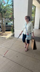 Woman with white cane, sunglasses, and shopping bag walking on city sidewalk.