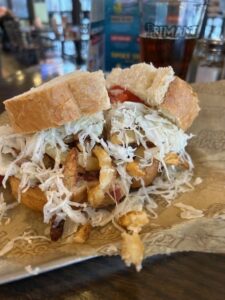 Delicious primanti-style sandwich with coleslaw, fries, and meat between thick slices of bread, sitting on a rustic wooden table in a bustling restaurant environment.