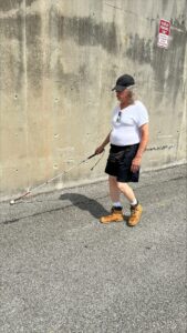 A man uses his white cane to trail a tall, cement wall on his right. He wears a black baseball cap, white t-shirt, and black shorts.
