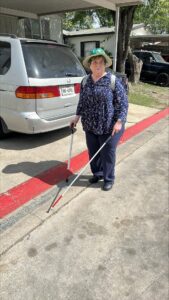 A women dressed in dark clothing and a wide-brimmed hat, uses a long cane in her left hand and a support cane in her right. She is following the red-painted curb on her right.