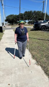 A woman using both a white cane and a support cane, wears dark clothing and a wide-brimmed hat while she walks down a sidewalk.