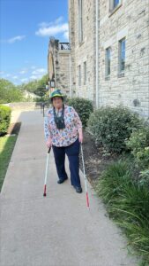 A woman walking around the outside a building built from limestone blocks. There is landscaping on either side of the sidewalk she is on, and she is using both a white cane and a support cane. 
