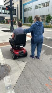 Two individuals waiting to cross the street, one in a red motorized wheelchair, the other providing support with a hand on their shoulder. Urban setting with modern buildings and a pedestrian signal showing a red hand.