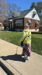 Person with a white cane walking towards a house on a sunny day. They are wearing a yellow Leader Dogs for the Blind hoodie, a plaid skirt, and sneakers.