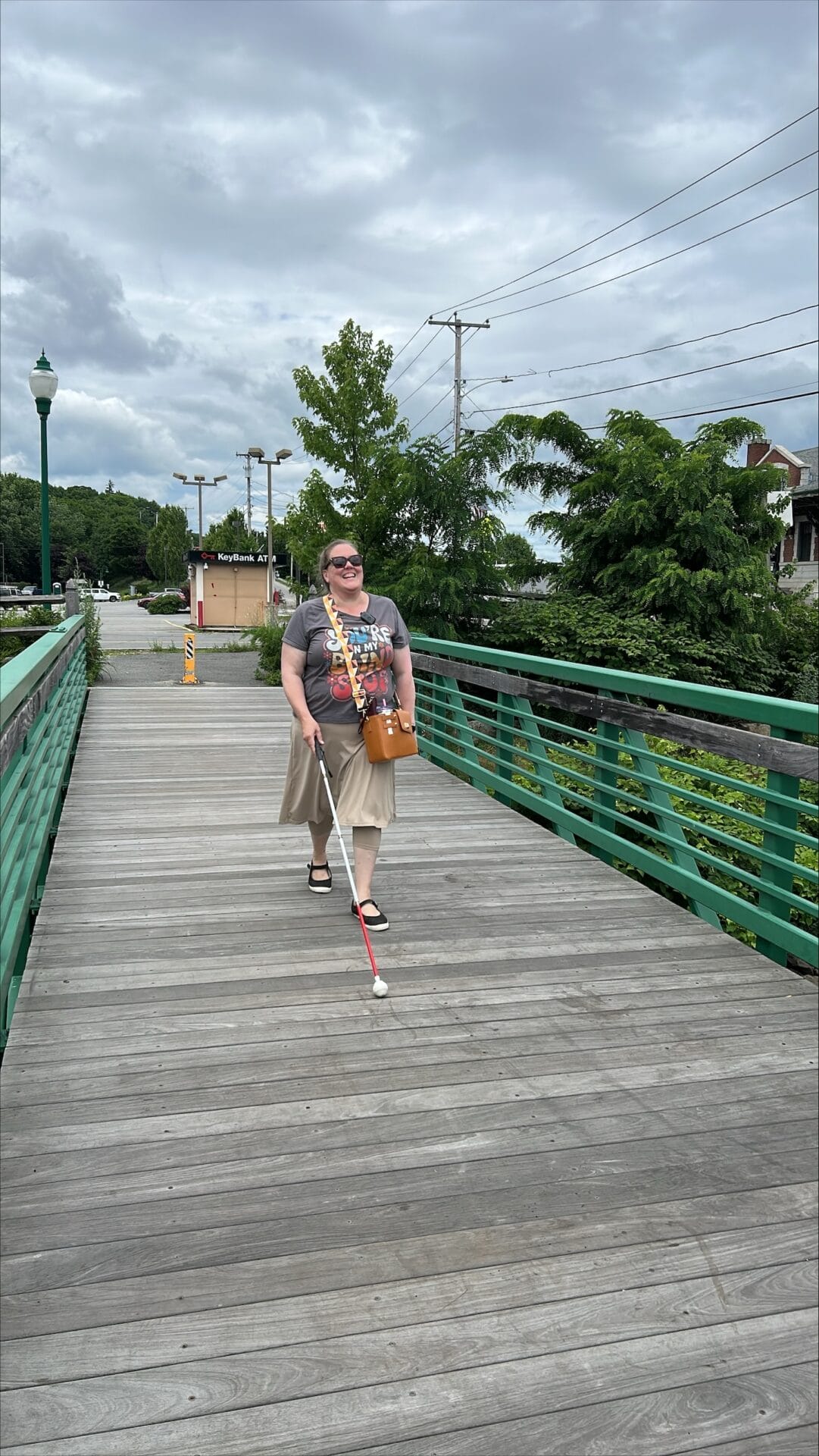 A person walks along a wooden bridge, using a white cane, surrounded by greenery and a cloudy sky.