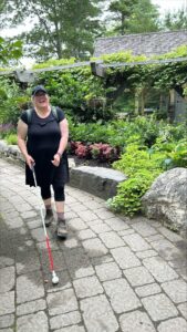A woman laughs as she uses her white cane to walk along a stone path. She is wearing all black and is surrounded by flora and fauna.