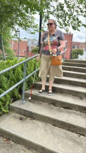 A woman uses her white cane to navigate stairs outdoors. She is wearing a t-shirt and a long skirt.