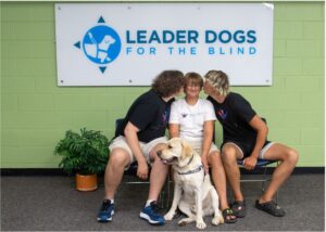 Three teenagers sit in front of a sign that reads “Leader Dogs for the Blind.” The two boys on either side are kissing the boy in the center on the cheek, making him squint his eyes. A yellow Labrador guide dog sits happily in front of them wearing a “Future Leader Dog” harness.