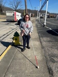 A woman wearing sunglasses walking down a sidewalk along a road with her white cane.