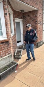 Person using a white cane walking past a brick house entrance.