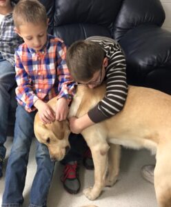 Two young boys sit on a bench petting and hugging a yellow Labrador. One boy wears a plaid shirt, and the other wears glasses and a striped shirt. The dog, standing between them, leans in affectionately while the boys interact with it warmly.