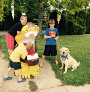 Three boys are playing near a colorful lion shaped play structure outdoors. One boy has his head inside the mouth of the lion, while another leans on it playfully, and the third stands smiling with a yellow Labrador wearing a blue vest labeled “Future Leader Dog.” The setting is a grassy area with trees and a sidewalk.