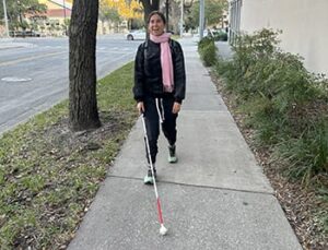 Smiling woman walking down a sidewalk toward the camera with a white cane. There is grass on either side of the sidewalk and some trees lining the sidewalk