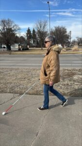 A person wearing a brown winter coat and sunglasses, travels down the sidewalk while using a white cane.