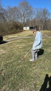 A person using a white cane in a grassy field with trees and a small building in the background.
