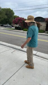 A man uses his white cane while walking down the sidewalk. He wears a straw hat, a blue, short-sleeved shirt, and long pants.
