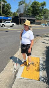 Holding his white cane upright, a man stands on bright yellow truncated domes on the street corner listening to traffic. He wears a straw hat, white t-shirt, and black shorts.
