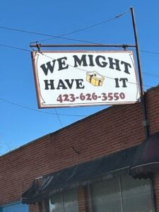 Sign reading "WE MIGHT HAVE IT" with a phone number, hanging outside a brick building under a clear blue sky.