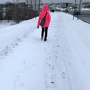 Person facing away from camera walking down side of snowy road with white cane. The person is weraing a bright pink/red coat and dark pants.