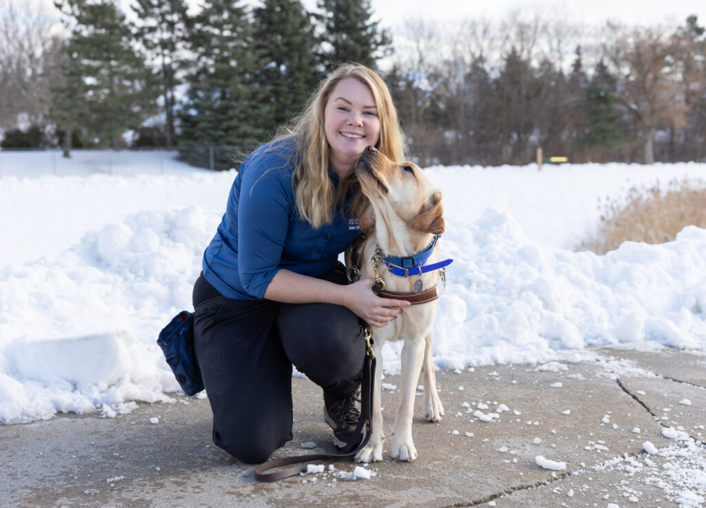 A woman with blond hair smiles toward the camera. Next to her is a yellow lab Leader Dog in harness sniffing at her face.