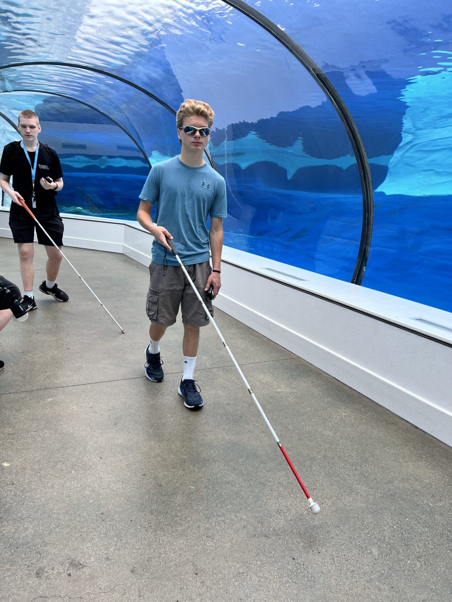 A person is walking through a tunnel at an aquarium, using a white cane for navigation while wearing sunglasses.