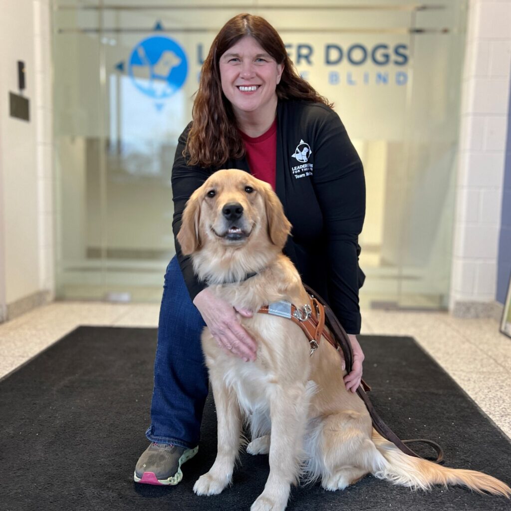 A woman with long brown hair kneels behind a golden retriever in the lobby of Leader Dogs for the Blind. The dog is wearing a leather guide dog harness.
