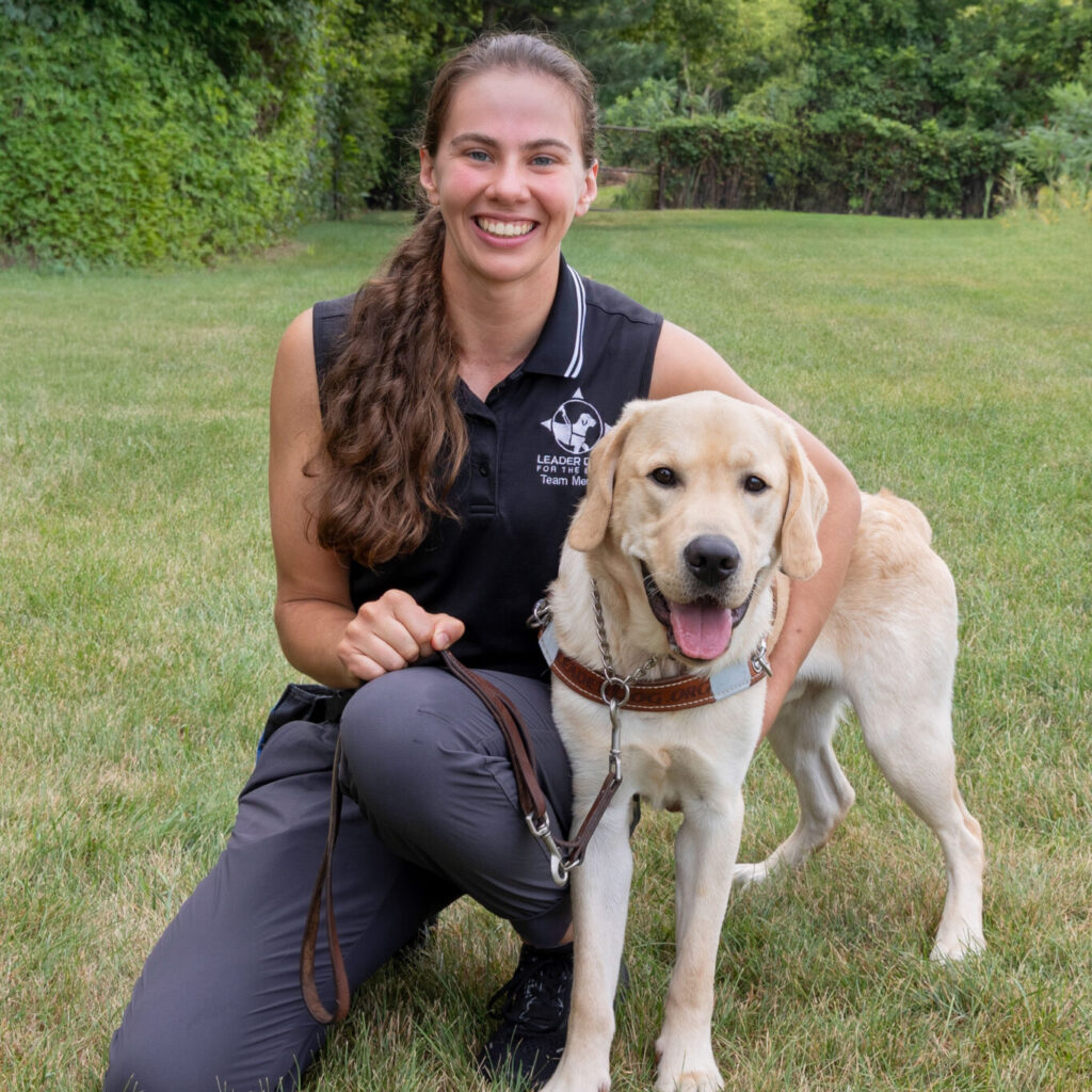 Person kneeling on a grassy lawn beside a yellow Labrador retriever wearing a guide-dog harness and leash, with trees and greenery in the background.