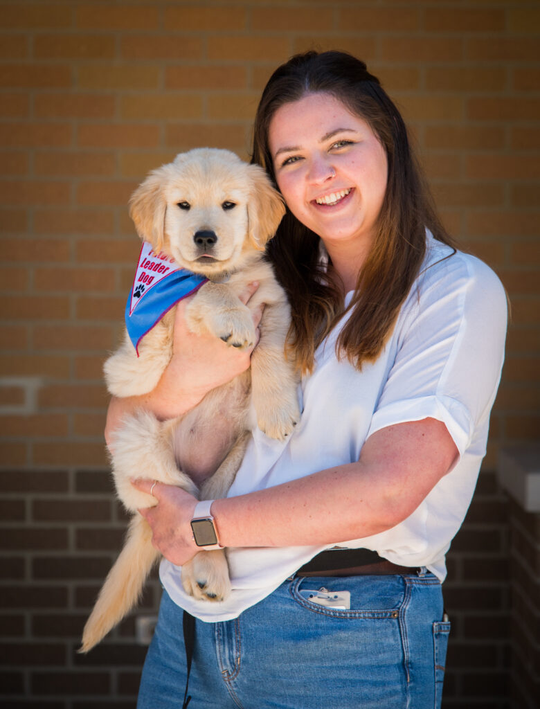 A woman holds a golden retriever puppy, both smiling against a brick background.