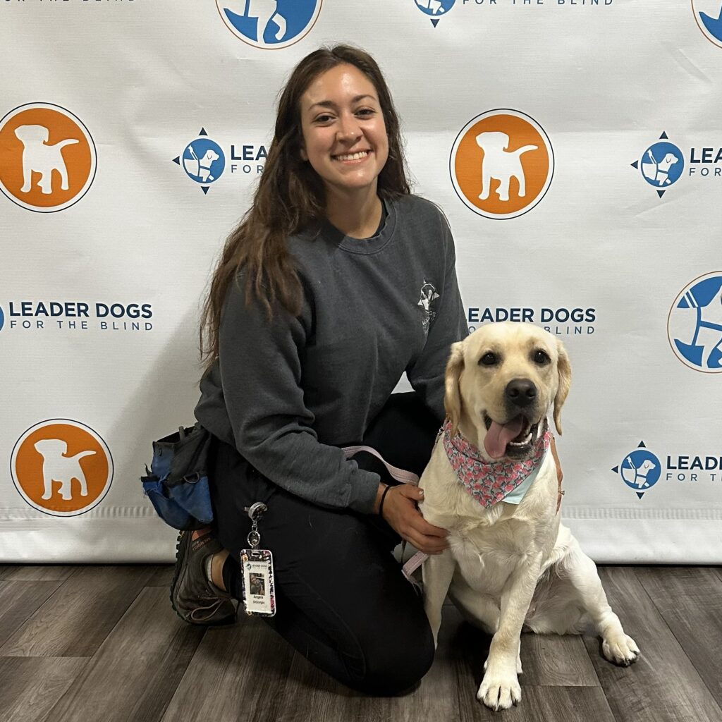 A woman with long brown hair kneels next to a yellow lab Leader Dog in harness. They both look toward the camera.