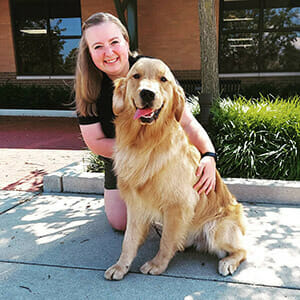 A person smiles while kneeling beside a golden retriever in a sunny outdoor setting.