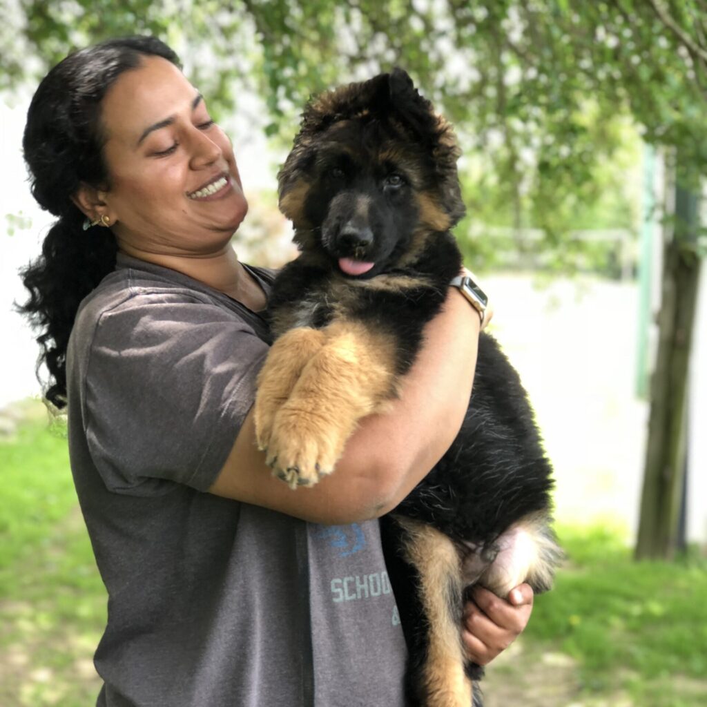 A person smiles widely while holding a black puppy close to their face.