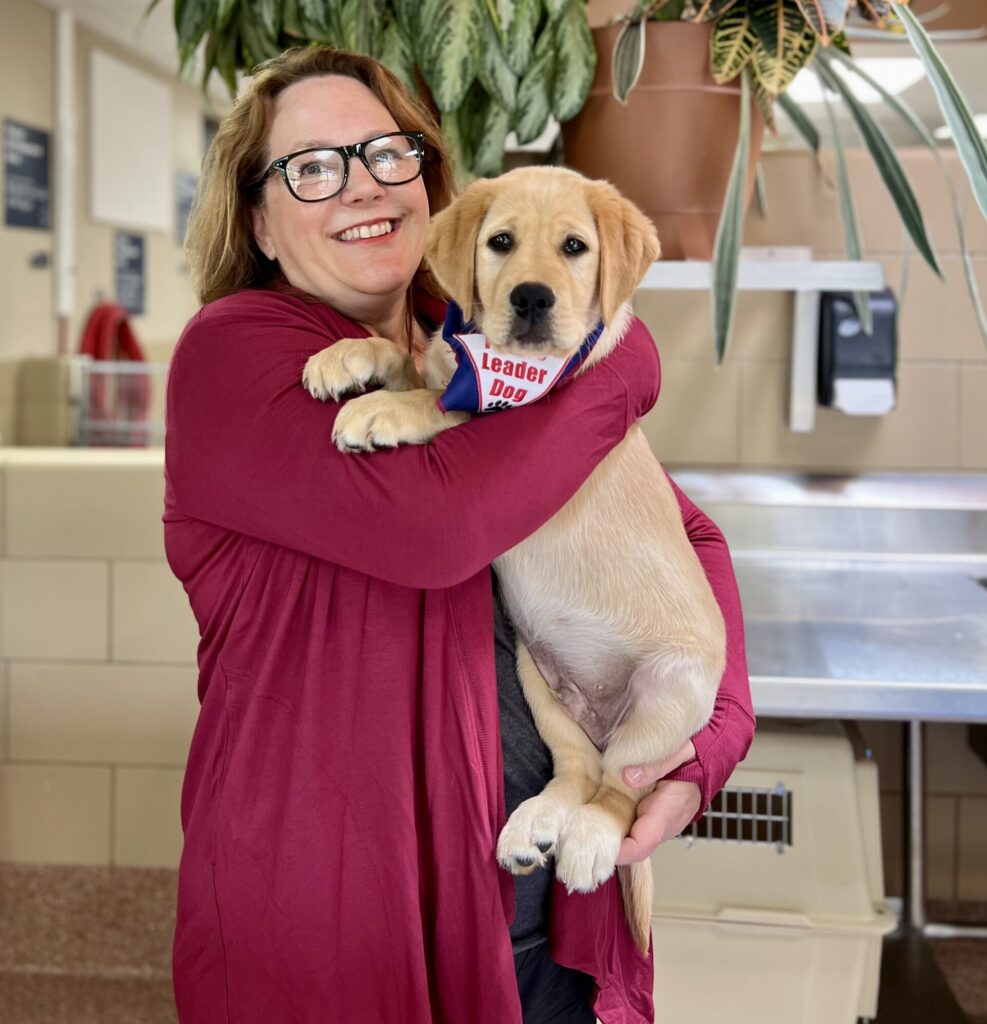 A woman in a maroon cardigan joyfully holds a golden Labrador puppy wearing a bandana that reads "Leader Dog."