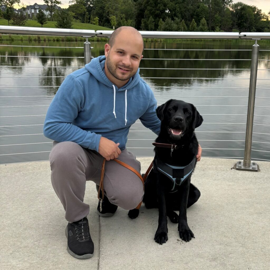 A man smiles toward the camera while kneeling next to a black lab Leader Dog in harness.