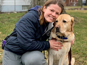 A person smiles while embracing a Labrador retriever, both sitting on the grass in a park-like setting.