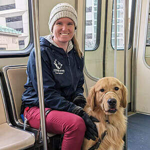 A person with a warm hat and gloves sits on a train seat next to a golden retriever.