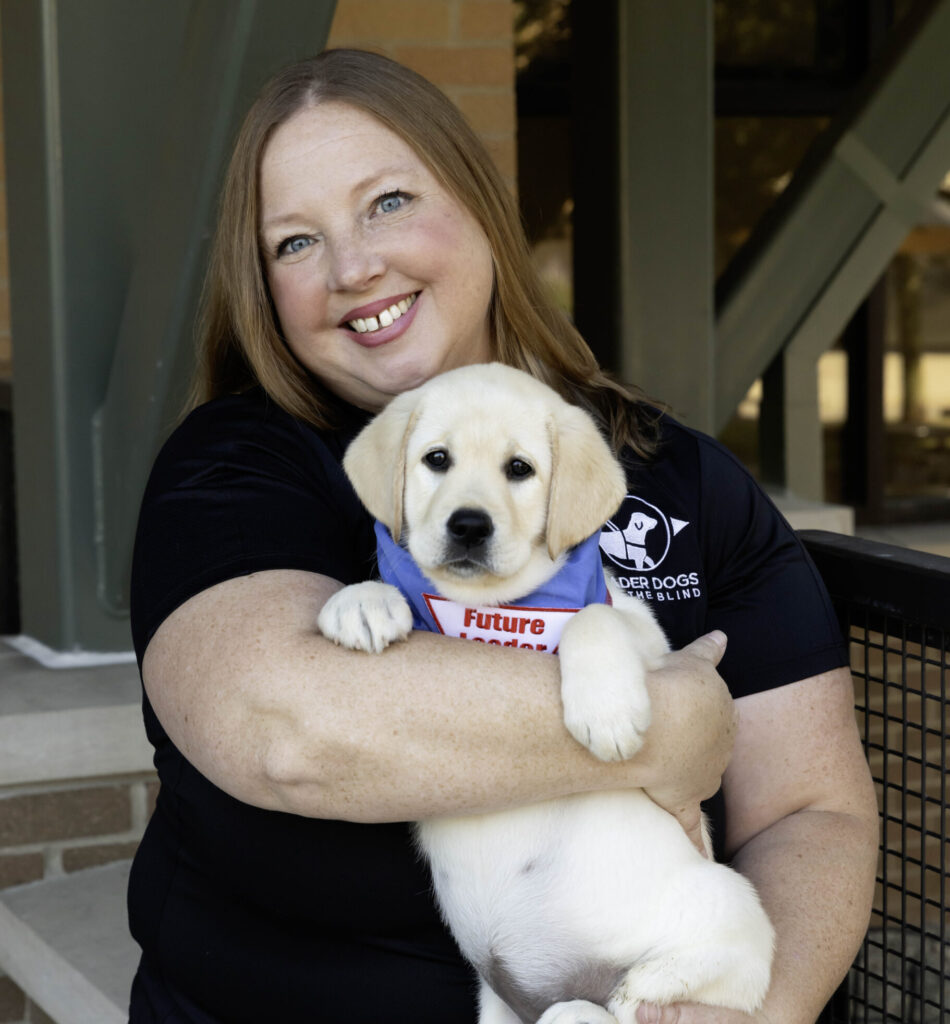 A smiling woman holds a fluffy puppy dressed in a blue vest, conveying warmth and connection.