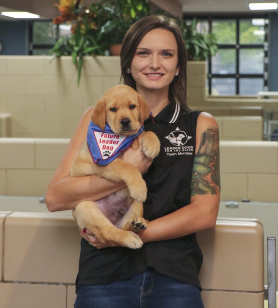 A person with a tattoo holds a puppy wearing a blue bandana that says "Future Leader Dog."
