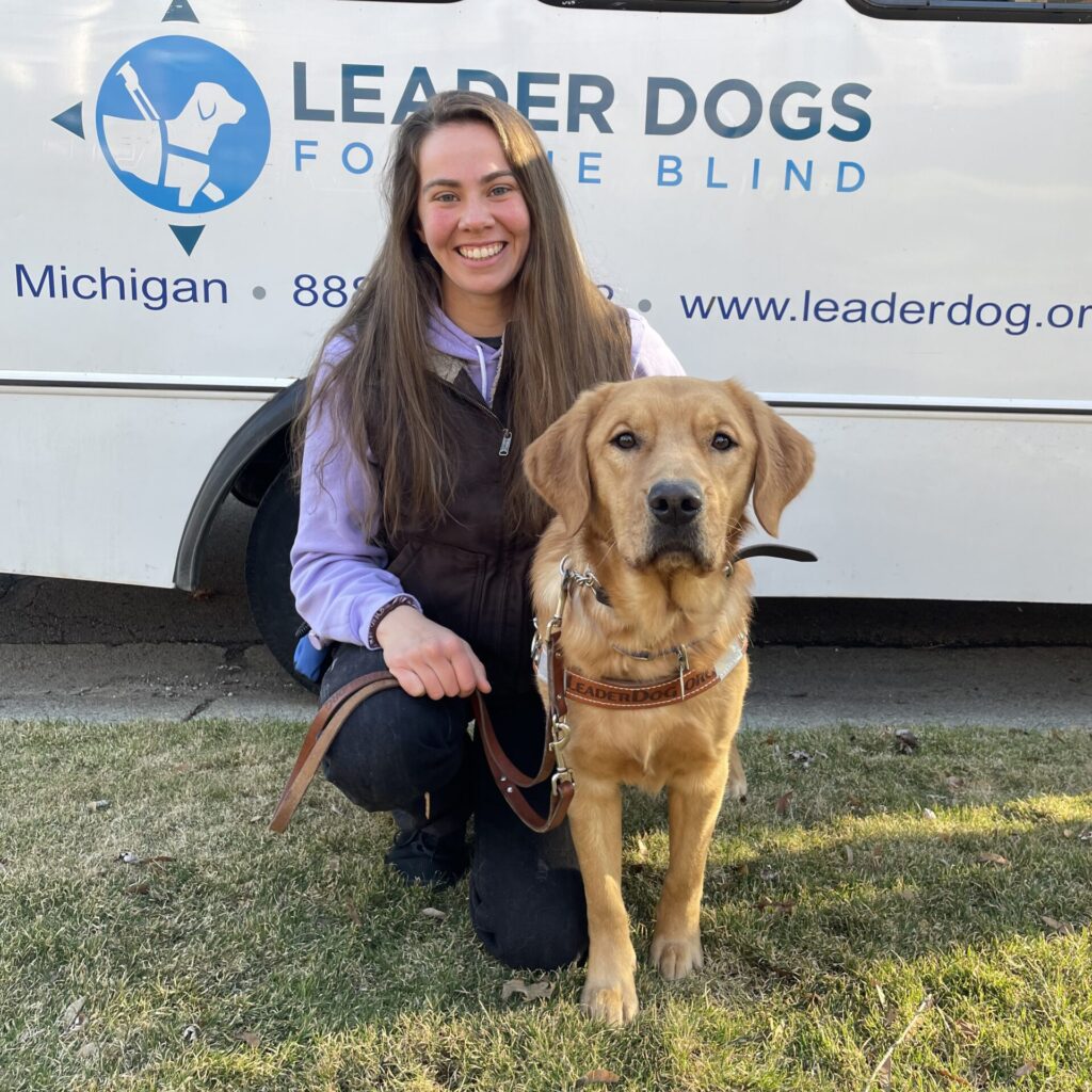 A person smiles while posing with a yellow Labrador retriever, both in front of a van associated with a guide dog organization.