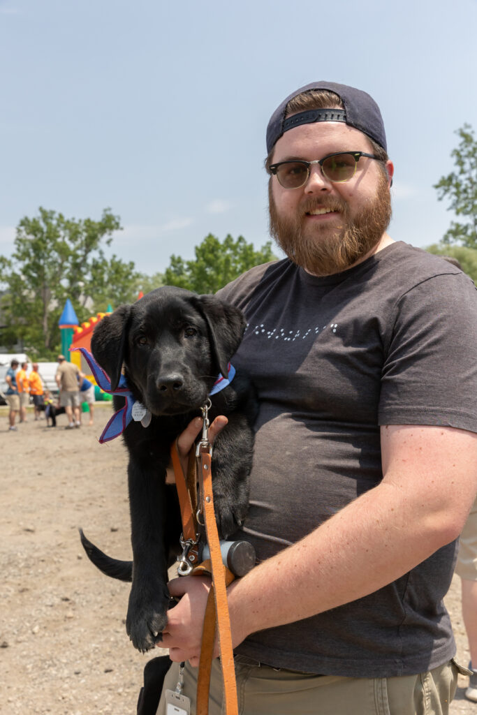 A man wearing glasses and a cap holds a cute black puppy with a colorful collar while standing outdoors.
