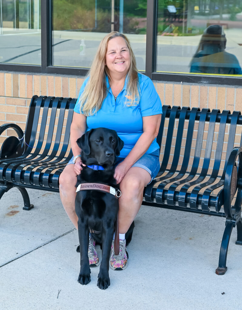 A woman in a blue shirt sits on a bench beside a black dog wearing a harness, smiling in a sunny outdoor setting.
