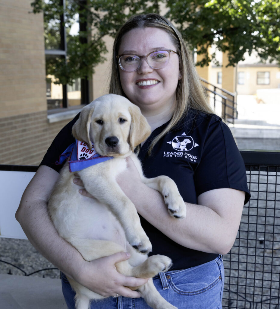 A smiling individual holds a playful golden Labrador puppy dressed in a blue bandana, both exuding warmth and joy in a bright outdoor setting.