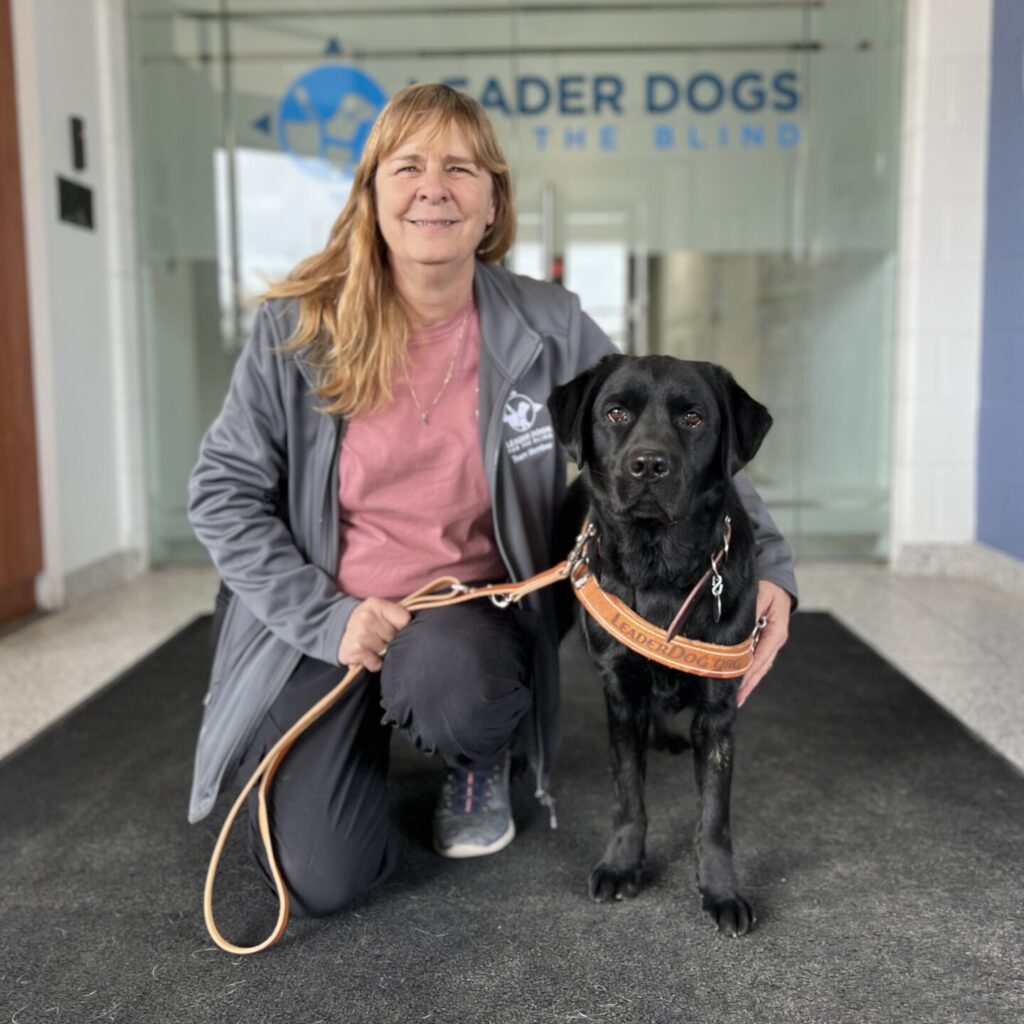 A woman smiles toward the camera while kneeling next to a black Lab Leader Dog in hist leather vest, The are in a hallway and the Leader Dogs for the Blind logo is visible behind them.