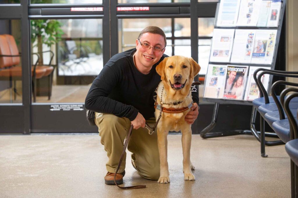 A smiling individual is kneeling next to a guide dog in a brightly lit indoor setting.