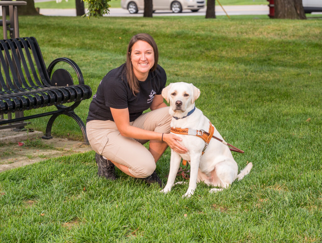 A person kneels beside a yellow Labrador wearing a harness in a grassy park setting.