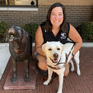 A smiling individual poses alongside a white dog and a bronze sculpture of a dog.