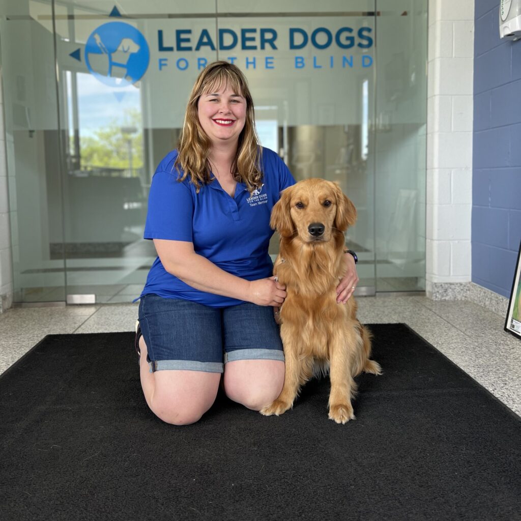 A person in a blue shirt smiles and crouches beside a golden retriever in front of a glass door with "LEADER DOGS FOR THE BLIND" displayed.