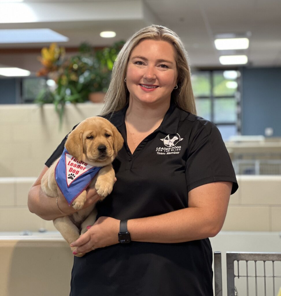 A woman in a black shirt holds a golden retriever puppy wearing a colorful bandana in a bright indoor setting.