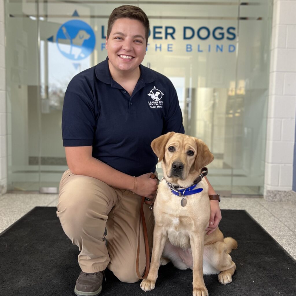A person in a navy polo shirt kneels beside a Labrador retriever, both smiling in an indoor setting with a sign for "Leader Dogs for the Blind" visible in the background.